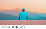A tranquil moment of contemplation as a man sits on a wall, admiring the expansive layered mountains bathed in the warm hues of a vibrant sunset sky. Стоковое AI-изображение, AI-художник Данила Большаков / Фотобанк Лори