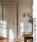 An elegant interior in Scandinavian or neoclassical style. A light wooden door with an expressive cornice and neat milling is combined with warm sunlight, softly falling on the walls and floor. A laconic wall lamp with a brass base and glass shades emphasizes the sophistication of the space. Comfort, silence and attention to detail make this scene ideal for interior and architectural projects, as well as advertising comfort and home style. Стоковое AI-изображение, AI-художник Анна Кривицкая / Фотобанк Лори