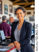Confident woman in a professional outfit stands at a reception desk in an automotive environment, smiling warmly while engaging with customers and showcasing a welcoming atmosphere. Стоковое AI-изображение, AI-художник Ирина Ручушкина / Фотобанк Лори