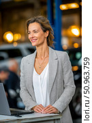 Confident woman in a stylish blazer stands beside a laptop in an automotive workshop, smiling warmly while engaging with clients in a professional environment. Стоковое AI-изображение, AI-художник Ирина Ручушкина / Фотобанк Лори