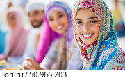 Close-up portrait of smiling woman wearing a colorful hijab, with group of people in modest attire blurred in the distance. Стоковое AI-изображение, AI-художник Данила Большаков / Фотобанк Лори