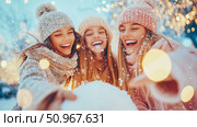 Three joyful friends enjoying a snowy winter outing with festive lights, embracing holiday spirit and laughter for a cheerful seasonal card or social media post. Стоковое AI-изображение, AI-художник Данила Большаков / Фотобанк Лори