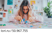 A focused young adult female with spectacles intently assembling a bright, multicolor jigsaw puzzle, ideal for educational game themes, mindful leisure, or concentration concepts. Стоковое AI-изображение, AI-художник Данила Большаков / Фотобанк Лори