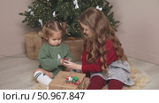 Two little girls, an older sister and a toddler, sit on the floor under the Christmas tree at home, playing with gifts and toys. Happy family and joy of winter holidays celebration concept. Footage. Стоковое видео, видеограф Ирина Михайличенко / Фотобанк Лори
