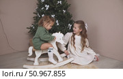 Two sisters have fun and play at home near the Christmas tree. The older sister sits on a floor, rocking a wooden rocking horse, while the younger toddler sister sits happily on it. Стоковое видео, видеограф Ирина Михайличенко / Фотобанк Лори