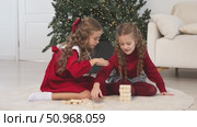 Happy family, little sister girls or best friends are sitting on floor near Xmas tree, playing board game with wooden tower together at home while spending leisure Christmas holidays. Footage. Стоковое видео, видеограф Ирина Михайличенко / Фотобанк Лори
