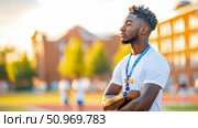 Profile portrait of a determined young Black man with dreadlocks and a whistle lanyard, standing on a sunlit sports field with his arms crossed, against a blurred university campus background. Стоковое AI-изображение, AI-художник Данила Большаков / Фотобанк Лори