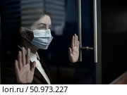 A sad quarantined woman in a business suit leaned against the window. Caucasian girl in a mask on isolation put her hands on a glass door. Maintaining social distance to prevent coronavirus. Стоковое фото, фотограф Михаил Решетников / Фотобанк Лори