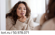 Woman applying lipstick while relaxing indoors on a cozy afternoon. Стоковое AI-изображение, AI-художник Katsiaryna Repich / Фотобанк Лори