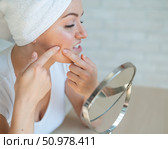 A woman with a white towel on head looks in the mirror and squeezes a pimple on cheek. A girl with hormonal problems does facial cleansing at home. Teen acne. Dermatologist himself in isolation. Стоковое фото, фотограф Михаил Решетников / Фотобанк Лори