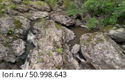 Aerial view of a serene waterfall cascading over rocky terrain surrounded by lush greenery, creating a tranquil natural scene with flowing water and textured stones. Стоковое видео, видеограф Ирина Ручушкина / Фотобанк Лори