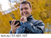 Portrait of mysterious attractive guy using gadget showing shh sign in park outdoors. Millennial man holding mobile phone doing silence gesture. Стоковое фото, фотограф Данила Большаков / Фотобанк Лори