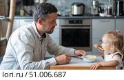 A caring man with a beard feeds his little daughter. A little girl sits at a children's dining table and dines with her dad. Happy fatherhood. Стоковое фото, фотограф Михаил Решетников / Фотобанк Лори