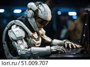 A futuristic cyborg in white armor with a high-tech helmet works at a computer in a dark high-tech environment. The image symbolizes the synergy of man and machine, artificial intelligence, technology development and cybersecurity. Ideal for topics related to the future of IT, robotics, science fiction, programming, hacking and virtual reality. The aesthetics are reminiscent of scenes from cyberpunk and sci-fi films. Стоковое AI-изображение, AI-художник Анна Кривицкая / Фотобанк Лори