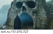 A dark-cloaked individual approaches a massive skull-shaped cave, surrounded by cliffs and misty ocean waters at dawn in a hauntingly beautiful setting. Стоковое AI-видео, аниматор Данила Большаков / Фотобанк Лори
