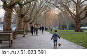 A homeless child playing with a ball in a public park, surrounded by winter trees, while adults walk by without noticing. Стоковое AI-изображение, AI-художник ALLA YURTAYEVA / Фотобанк Лори