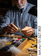 Senior fly tier crafting artificial fishing lures at workshop desk. Стоковое AI-изображение, AI-художник Юрий Поздников / Фотобанк Лори