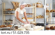 Professional young female baker in white uniform kneading dough on floury table in artisan bakery shop, surrounded by freshly baked bread and pastries arranged on wooden shelves. Стоковое видео, видеограф Яков Филимонов / Фотобанк Лори