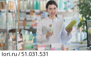 Young woman pharmacist holding medicated shampoos for hair treatment in her hands and standing near counter with computer in pharmacy. Стоковое видео, видеограф Яков Филимонов / Фотобанк Лори