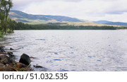 Lake Bannoe, Russian Federation - July 31, 2025: Serene lakeside view with gentle waves lapping against rocky shore, surrounded by lush greenery and distant mountains under a cloudy sky. Редакционное видео, видеограф Ирина Ручушкина / Фотобанк Лори
