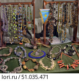 An exhibition stand with a collection of jewelry made of natural stones. Beads and necklaces made of amethyst, turquoise, amber, agate and other minerals are hung on wooden stands. Handmade bracelets, necklaces and pendants are laid out on the table. The jewelry is distinguished by a variety of shapes, colors and styles, creating a bright and rich composition. Стоковое фото, фотограф Анна Кривицкая / Фотобанк Лори