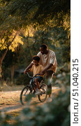 Father teaches son to ride bike in sunlit park during late afternoon, enjoying quality time together outdoors. Стоковое AI-изображение, AI-художник Сергей Толмачёв / Фотобанк Лори