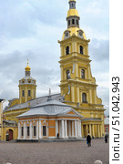 A general view of the Peter and Paul Cathedral in St. Petersburg, located on the territory of the Peter and Paul Fortress. In the foreground is a chapel with a colonnade and a triangular pediment, painted in warm orange and white tones. Behind it rises a majestic bell tower with a golden spire topped by an angel and a clock on the facade. On the right is the main dome of the cathedral with a golden cross and decorative elements in the Baroque style. The stone-paved square in front of the building gives the frame an atmosphere of old St. Petersburg. The weather is cloudy, which emphasizes the contrast between the gray sky and the bright yellow and gold facades. (2013 год). Редакционное фото, фотограф Анна Кривицкая / Фотобанк Лори