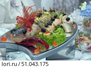 An exquisite festive dish — stuffed and decorated fish served on a large mirrored platter. The head of the fish is decorated with an edible element of bright red caramelized sugar, and the eyes are decorated with olives. Around the fish are pieces of fillet, lemon rolls, olives on skewers, fresh herbs, lettuce leaves and red caviar. In front, near the head of the fish, lies a boiled bright red crayfish, adding to the effect of the composition. The garnish and decor are arranged symmetrically, creating a festive and luxurious look. The dish is served at a banquet, as evidenced by the glasses, salads and smartly dressed guests in the background. Стоковое фото, фотограф Анна Кривицкая / Фотобанк Лори