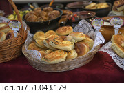 An appetizing assortment of fresh baked goods laid out in wicker baskets on a table covered with a burgundy patterned tablecloth. In the center of the frame are ruddy buns and pies of various shapes: puff pastry curls with poppy seeds, soft pastries with a golden crust, neatly stacked. On the left and right in the frame you can see baskets with other types of baked goods - cheesecakes with berries, cherry pies, envelopes with ham and cheese. White paper napkins with an openwork edge emphasize the neat presentation. The background is slightly blurred, but you can make out ceramic pots and other food, which creates an atmosphere of home comfort and a village fair. The photo conveys warmth, the aroma of fresh bread and a welcoming atmosphere. Стоковое фото, фотограф Анна Кривицкая / Фотобанк Лори