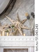 Light interior with elements of winter and New Year's decor. In the foreground is a snow-white fireplace with brickwork and a decorative carved cornice. On the mantelpiece are cream cylindrical candles of different heights, an elegant New Year's composition with fir branches, cones, silver balls and a bow made of silver ribbon. On the wall behind the fireplace are two large paper stars with an openwork ornament in warm beige and gray tones. On the right, in a glass vase, there are decorative balls on long stems in silver and cream colors. The background of the wall is made of gray and white textured decorative plaster. The atmosphere of the photo is warm and cozy, in the Scandinavian style, with elements of minimalism and festive decoration. Стоковое фото, фотограф Анна Кривицкая / Фотобанк Лори