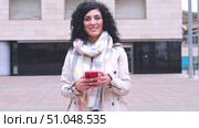 Confident woman with scarf holding smartphone and smiling at camera in the city. Стоковое видео, видеограф Raquel Corcoles Mendez / Фотобанк Лори