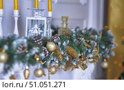 New Year's composition for interior decoration. On the white mantelpiece there is a garland of artificial pine with snow-covered branches, cones, golden balls and decorative elements. The decorations are complemented by golden cones, beads and figurines, creating an atmosphere of luxury and celebration. In the background you can see a white metal lantern with openwork patterns and yellow candles in tall white candlesticks. Soft bokeh in the background gives the composition coziness and warmth, and golden accents harmoniously combine with green and white colors. Стоковое фото, фотограф Анна Кривицкая / Фотобанк Лори