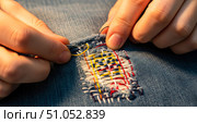 Hands visible mending torn blue jeans with colorful threads, closeup of person repairing clothes with needle and thread using sashiko technique. Стоковое AI-изображение, AI-художник Павел Муравьев / Фотобанк Лори