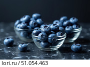 Plump blueberries are beautifully arranged in three glass bowls atop a dark marble countertop, showcasing their vibrant color and inviting texture. Perfect for snacking or baking. Стоковое AI-изображение, AI-художник Ольга Игнашина / Фотобанк Лори