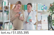 Interested pregnant woman using smartphone to check ingredients of organic shampoo at drugstore, while smiling professional female pharmacist in background suggesting different pregnancy-safe option. Стоковое видео, видеограф Яков Филимонов / Фотобанк Лори