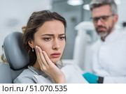 Красивая молодая женщина сидит в современном стоматологическом кресле, приложив руку к щеке от зубной боли, с рядом стоящим мужчиной-стоматологом в белом халате. Стоковое AI-изображение, AI-художник Максим Коломыченко / Фотобанк Лори
