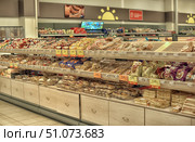 A view of the bakery section of a supermarket. The shelves are neatly lined with packaged products — bread, rolls, crackers, cookies, and various types of baked goods. The products are arranged in several rows, each with its own price tag, including discounted offers. The shelves below are lined with packages of bread and rolls, and above are a variety of cookies and sweets in colorful packages. Other sections of the store are visible in the background, and the walls are decorated with images of the sun. The lighting is warm, creating a cozy atmosphere, emphasizing the freshness and attractiveness of the products. (2015 год). Редакционное фото, фотограф Анна Кривицкая / Фотобанк Лори