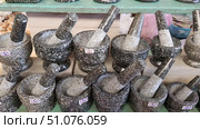A row of granite mortars and pestles displayed on a shelf for sale. Each mortar is made of a durable grey stone with a distinctive marble pattern for durability and stability. The pieces vary in size and price, indicated on stickers in Thai baht. The larger mortars are in the foreground, while the smaller ones are in the background. The background includes elements of a marketplace: wooden shelves, baskets and packaging. These kitchen utensils are traditionally used for grinding spices, herbs and nuts, as well as for making pastes and sauces. Стоковое фото, фотограф Анна Кривицкая / Фотобанк Лори