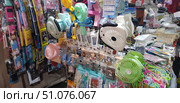 A bright and colourful market stall with a variety of goods. In the foreground hang compact portable fans of various colours – green, pink, mint, white and black. The display case also features mobile phone cases with cartoon characters, keychains, smartphone holders, cables, and at the bottom – multi-coloured laces and accessories. In the background is a table with various small goods: tools, packaging, boxes, gadgets. The atmosphere of a lively street trade, an abundance of small items and bright details. (2018 год). Редакционное фото, фотограф Анна Кривицкая / Фотобанк Лори