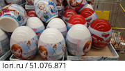 A close-up of a shop window or product display. In the foreground are large plastic eggs with images of Disney and Pixar characters. The blue and white eggs are Frozen-themed with images of Anna, Elsa, and Olaf. The red and white eggs are Cars-themed with images of Lightning McQueen. These eggs usually contain sweets and toys, which attracts children. The products are neatly packed into boxes, some of which are in a trolley. The background is the retail space of the shop. (2018 год). Редакционное фото, фотограф Анна Кривицкая / Фотобанк Лори