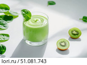 A velvety green smoothie rests in a sleek glass, atop a pristine white surface. Surrounded by fresh spinach, cucumber, and kiwi, it offers a minimalist yet vibrant display of nutritious ingredients. Стоковое AI-изображение, AI-художник Nart / Фотобанк Лори