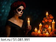 A woman with skull makeup and a flower crown stands beside a candlelit altar adorned with marigolds, celebrating Día de los Muertos. Стоковое AI-изображение, AI-художник Nart / Фотобанк Лори