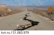 A cracked road stretches through a barren landscape. Mountains are visible in the background. A warning sign stands beside the damaged pavement. Стоковое AI-видео, аниматор Nart / Фотобанк Лори