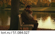 A man with dark hair and beard sits on a bench by a lake, holding a young boy with light brown hair. Autumn leaves surround them, creating a warm atmosphere. Стоковое AI-видео, аниматор Nart / Фотобанк Лори