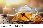 A glass teapot filled with herbal tea sits on a wooden table. Slices of orange, cinnamon sticks, and star anise surround the teapot. A candle glows softly in the background. Стоковое AI-видео, аниматор Nart / Фотобанк Лори