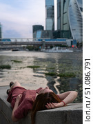 Young woman in a burgundy dress relaxing on a stone embankment, lying with her head back and hand resting on her forehead near the water. Стоковое фото, фотограф Софья Соломонова / Фотобанк Лори
