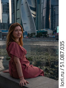 Woman in a burgundy dress sits on the embankment with skyscrapers of Moscow City in the background. Редакционное фото, фотограф Софья Соломонова / Фотобанк Лори