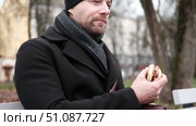 Pensive hipster drinking coffee sitting on a bench in the urban park. Handsome man wearing black coat eating street food, hot dog with paper cup in hand in the city. Stylish bearded middle-aged male. Стоковое видео, видеограф Марина Демидюк / Фотобанк Лори