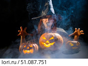 An ominous witch in a hat conjures over a jack-o-lantern. Traditional halloween characters. Mystical fog creeps over pumpkins with carved terrible faces. An evil witch casts a spell. Стоковое фото, фотограф Михаил Решетников / Фотобанк Лори