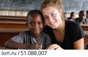 European smiling teacher teaching students in rural African school. Black African children from disadvantaged families study in school, read books, receive primary education, volunteer service. Стоковое AI-изображение, AI-художник ALLA YURTAYEVA / Фотобанк Лори